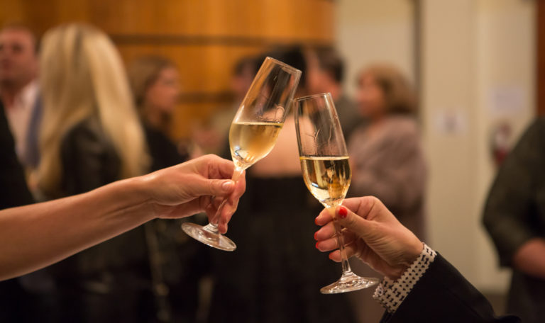 2 hands clinking together champagne glasses with guests socializing in the background.