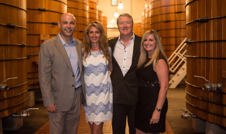 2 women and 2 men in cocktail attire smiling in front of gigantic tun barrels of wine in Jordan Winery's cellars.