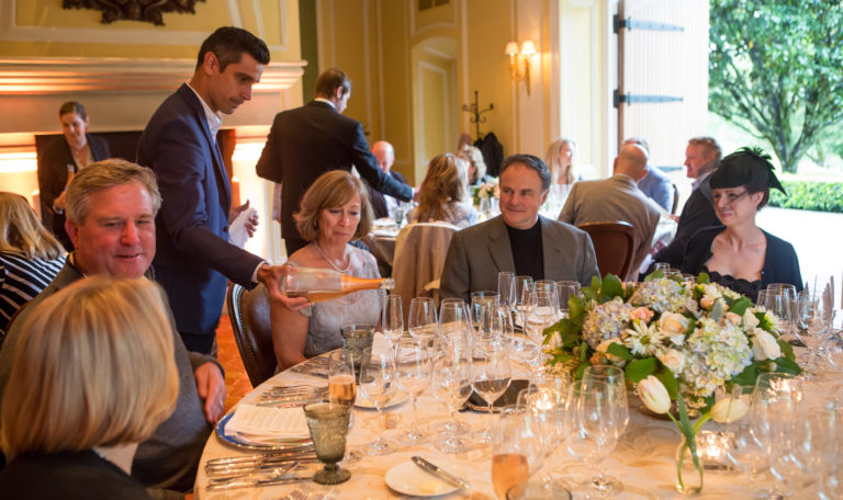 A waiter pouring champagne for a party of guests in formal attire at a round table set with various tasting glasses.