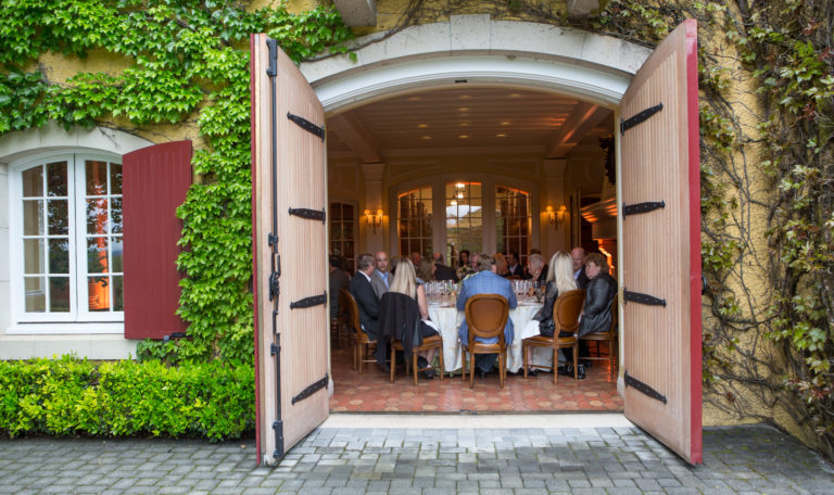 Looking into Jordan Winery's tasting room through large wooden double doors. Guests in formal attire are seated inside. The exterior walls of the building are covered in lush ivy and vines.