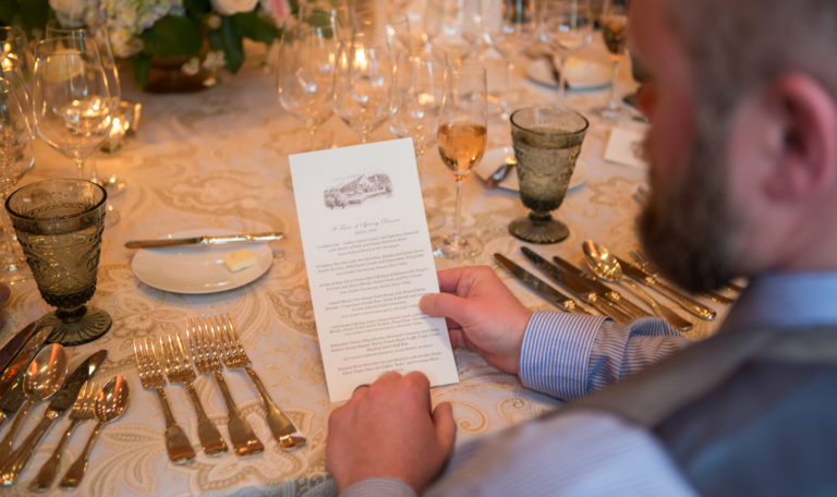 Looking over a man's shoulder as he holds Jordan Winery's Spring Dinner tasting menu at a formally set table.
