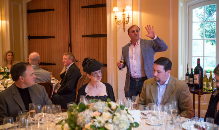 A man in a white button down and grey blazer toasts a tasting room full of guests. Behind him to the left are large wooden double doors.