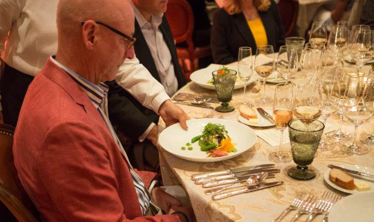 A waiter placing a dish in front of a man in a dusty red blazer seated at a formally set tasting table.