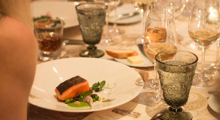 Looking over a woman's shoulder at the salmon dish placed at a formally set tasting table. Glasses are filled with chardonnay and champagne.