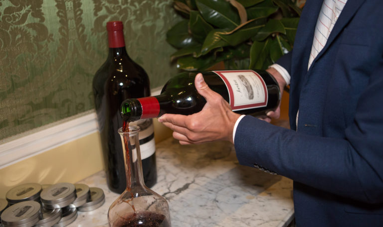 A man in a navy blue suit pours a Double Magnum bottle of Jordan's Cabernet Sauvignon into a decanter. On the white marble table is another large bottle of red wine.