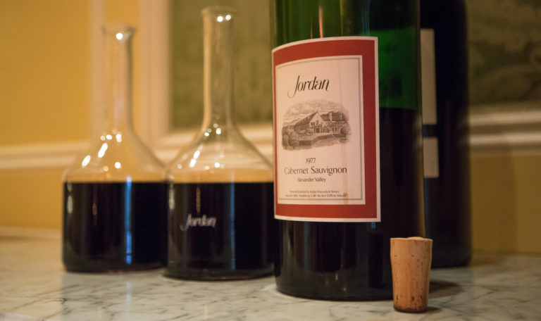 Close-up of 2 decanters of red wine next to a Double Magnum bottle of Jordan's 1977 Cabernet Sauvignon and a cork on a white marble table.