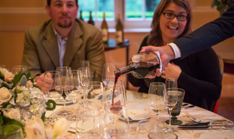 A waiter's right hand pours red wine from a decanter into a glass on a formally set table. A woman and man look on smiling.