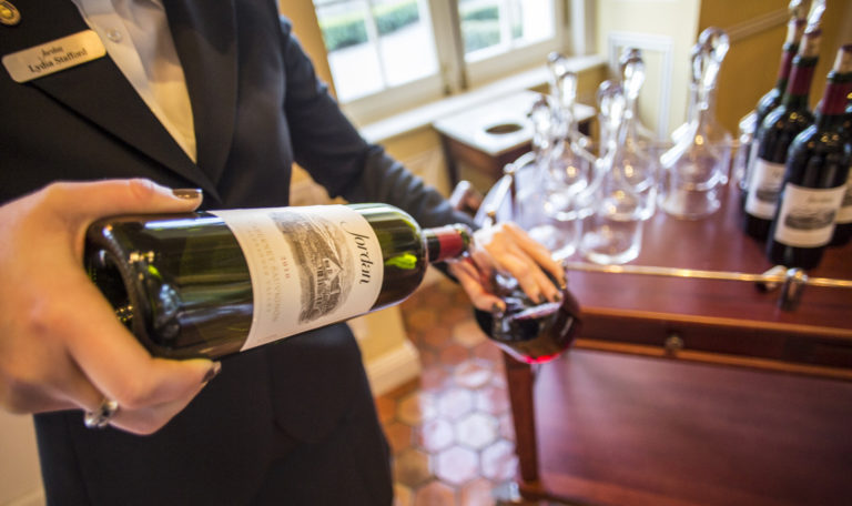 A waiter's hands pouring Jordan's Cabernet Sauvignon into a decanter next to a wooden bar cart. The perspective is a high angle looking down the bottle.