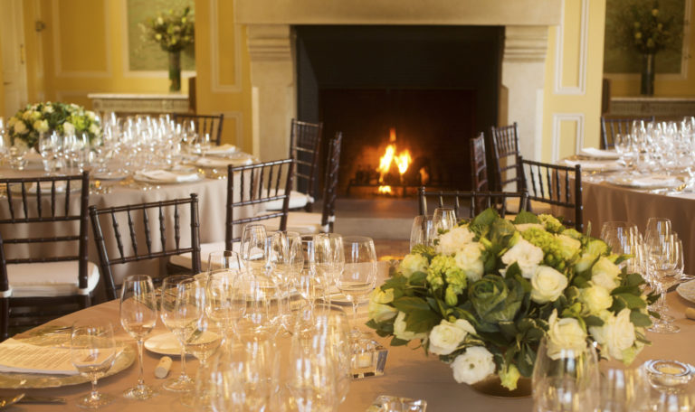 3 formally set round tasting tables in front of a white column fireplace. White rose bouquets sit centered on beige table cloths.