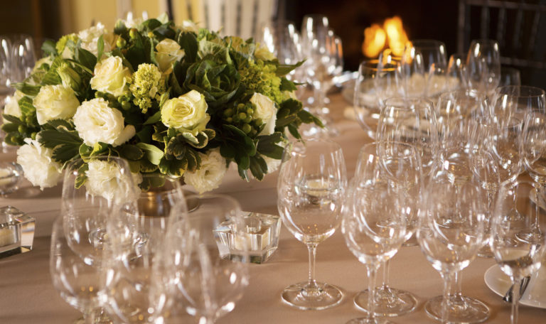 Multiple empty wine glasses on a beige table cloth next to a white rose bouquet.