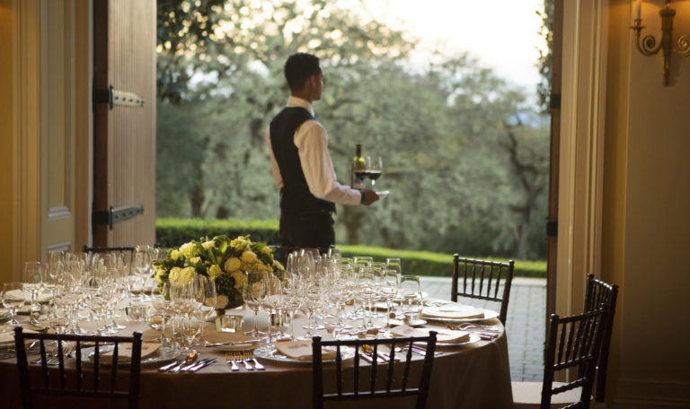 A round formally set tasting table next to large open wooden double doors. Centered is a waiter exiting to serve 2 glasses of red wine.