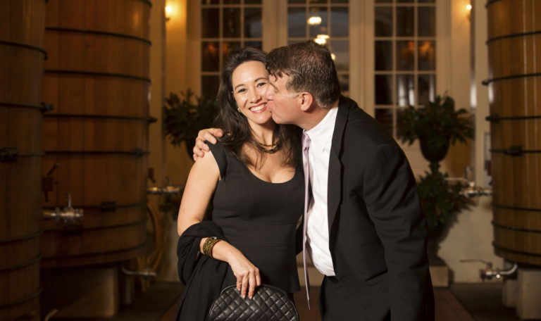 A woman with black hair and a black dress smiles into the camera as a man in a suit and pink tie kisses her left cheek. They are standing in front of gigantic tun barrels of wine.