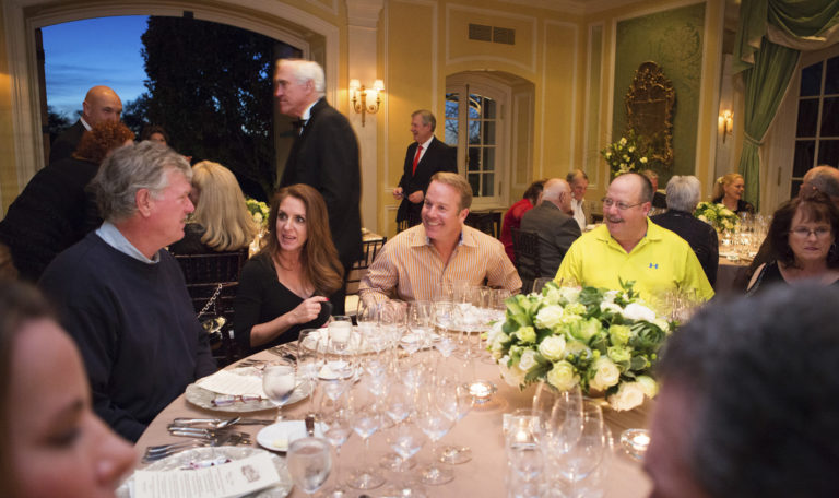 Guests seated at a beige circular dinner table with formal settings at Jordan Winery's Valentine's dinner event. 2 men on the right smile at another man across the table. The woman between appears to be telling them all a story. Men in black suits cross in the background.