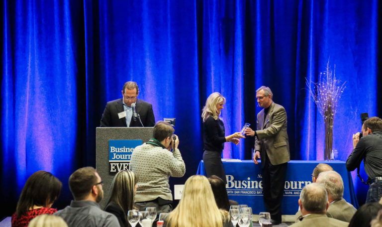 A blonde woman accepting the North Bay Business Journal Award from a man in a grey blazer on stage in front of photographers. Another man is preparing to read a speech at the podium. Blue velvet curtains are the backdrop.
