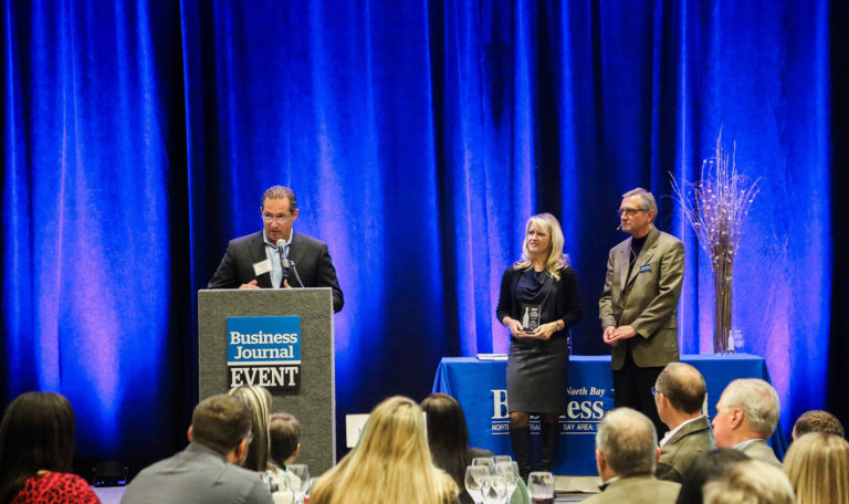 A man at a podium reading an acceptance speech for the North Bay Business Journal Award on stage. On the right stands a blonde woman holding the glass trophy next to a man in a grey blazer. Blue velvet curtains are the backdrop.
