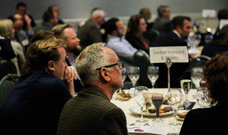 Right profile view of guests sitting at an event table for the North Bay Business Journal Awards. Centered is a man with white peppered hair and glasses. On the table a sign reads "Jordan Vineyard & Winery."