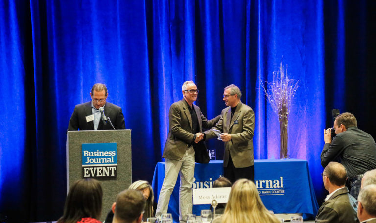 2 men in grey blazers shake hands on stage in front of photographers as 1 accepts the North Bay Business Journal Award. On the left a man prepares to read at the podium. Blue velvet curtains are the backdrop.