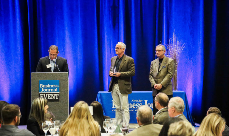 On the left a man at a podium reads a speech as 2 other men stand on the right. The center man holds the North Bay Business Journal Award. Blue velvet curtains are the backdrop.