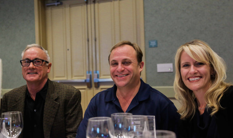 A man with white hair and glasses, a man in a navy blue polo, and a blonde woman smile at the camera. They are attending the North Bay Business Journal Awards. Behind them are tan double doors.