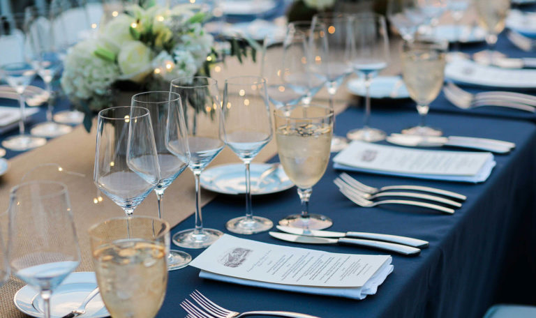 Close-up of Wine Dinner Starlight Supper table settings each with 4 empty wine glasses, a narrow menu, and a goblet of water. There's a navy blue table cloth with a burlap runner. Small white bouquets line the center.