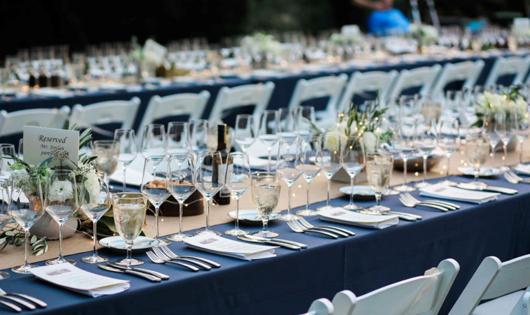 Wine Dinner Starlight Supper table settings each with 4 empty wine glasses, a narrow menu, and a goblet of water. There's a navy blue table cloth with a burlap runner. Small white bouquets line the center. In the background is an identical table.