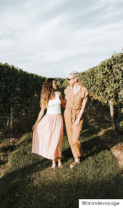 2 women smiling at each other in a grassy vineyard row. The left woman has a white tank top and long pink striped skirt. The right woman has a caramel jumpsuit with cinched waist belt. Both have sunglasses.