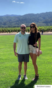 A couple standing on a lawn in front of a vineyard. He wears a pastel green polo and grey shorts. She has a black shirt and white plaid shorts. Both wear sunglasses.