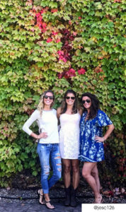 3 women posing in front of an ivy wall. The left woman has ripped jeans and long sleeve white shirt. The center woman has a short white lace floral dress and black cowgirl boots. The right woman has a blue floral t-shirt dress. All wear sunglasses.