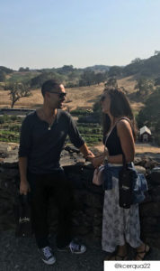 A couple leaning against a stone wall smiling at each other overlooking a garden. He wears a gray henley shirt, dark jeans, and converse. She has a black tank top and long white floral skirt.