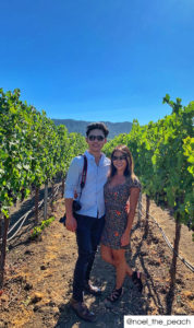 A couple smiling in a lush vineyard row. He wears a button down, jeans, and nice boots. She has a short black and white dress with small orange flowers.