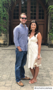 A couple standing outside in front of large arched wooden double doors. He wears a navy gingham plaid button down and jeans. She has an eggshell knee length dress with buttons down the front.