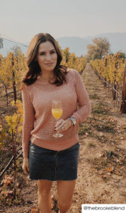 Woman standing in vineyard in fall holding a mimosa. She's wearing a peach open knit sweater and short black denim skirt.