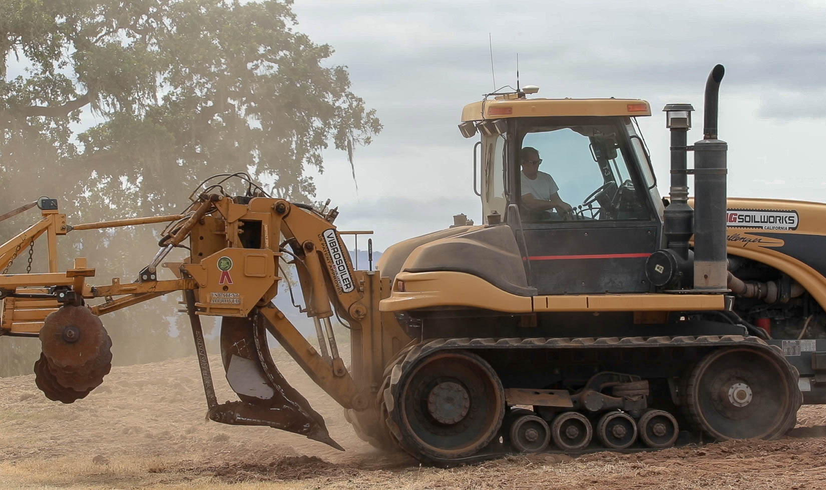 Profile of a vineyard soil deep ripping tractor. Dust hangs in the air and a man in a white t-shirt and sunglasses sits in the cab.