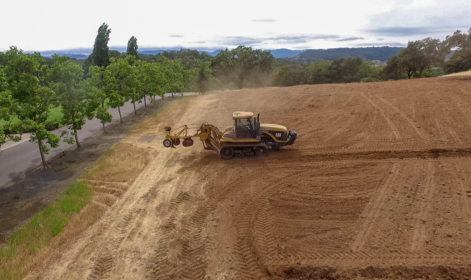 Yellow vineyard soil deep ripping tractor in a freshly tilled dirt field. Trees line the street on the left.