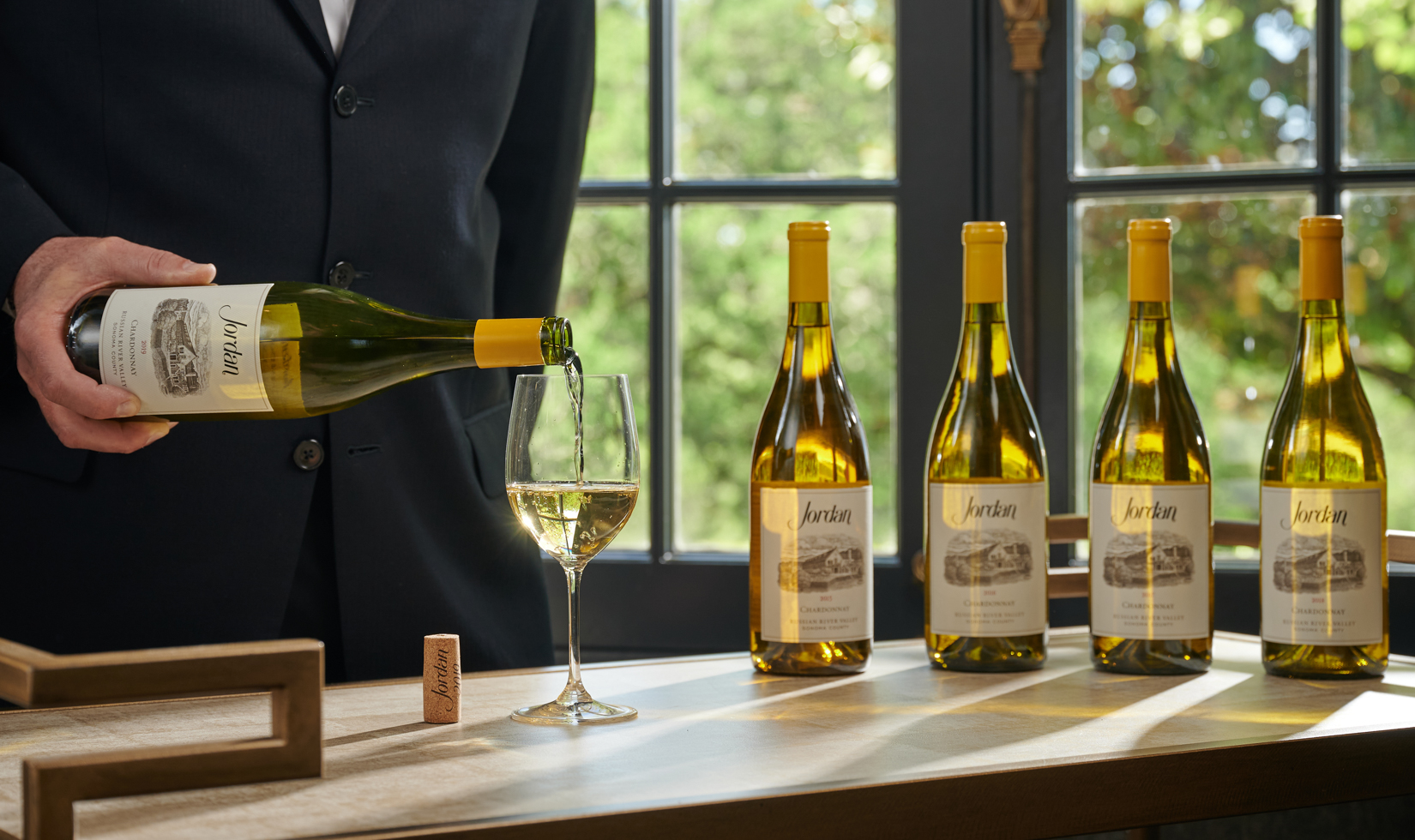 chardonnay wine pouring into a wine glass with four wine bottles behind it on a table near window