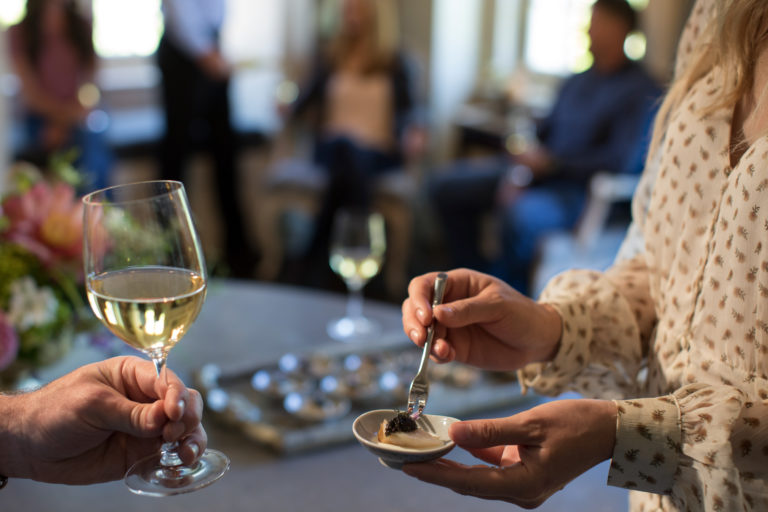 Hand holding a glass of white wine while another person holds an appetizer