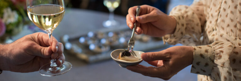 Hand holding a glass of white wine while another person holds an appetizer