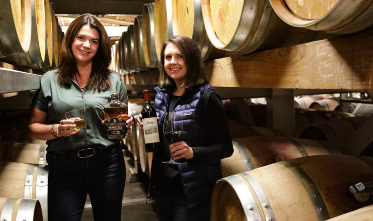 Woman in green shirt and jeans holding whiskey bottle on the left with woman in black shirt and jeans holding a cabernet bottle on the right