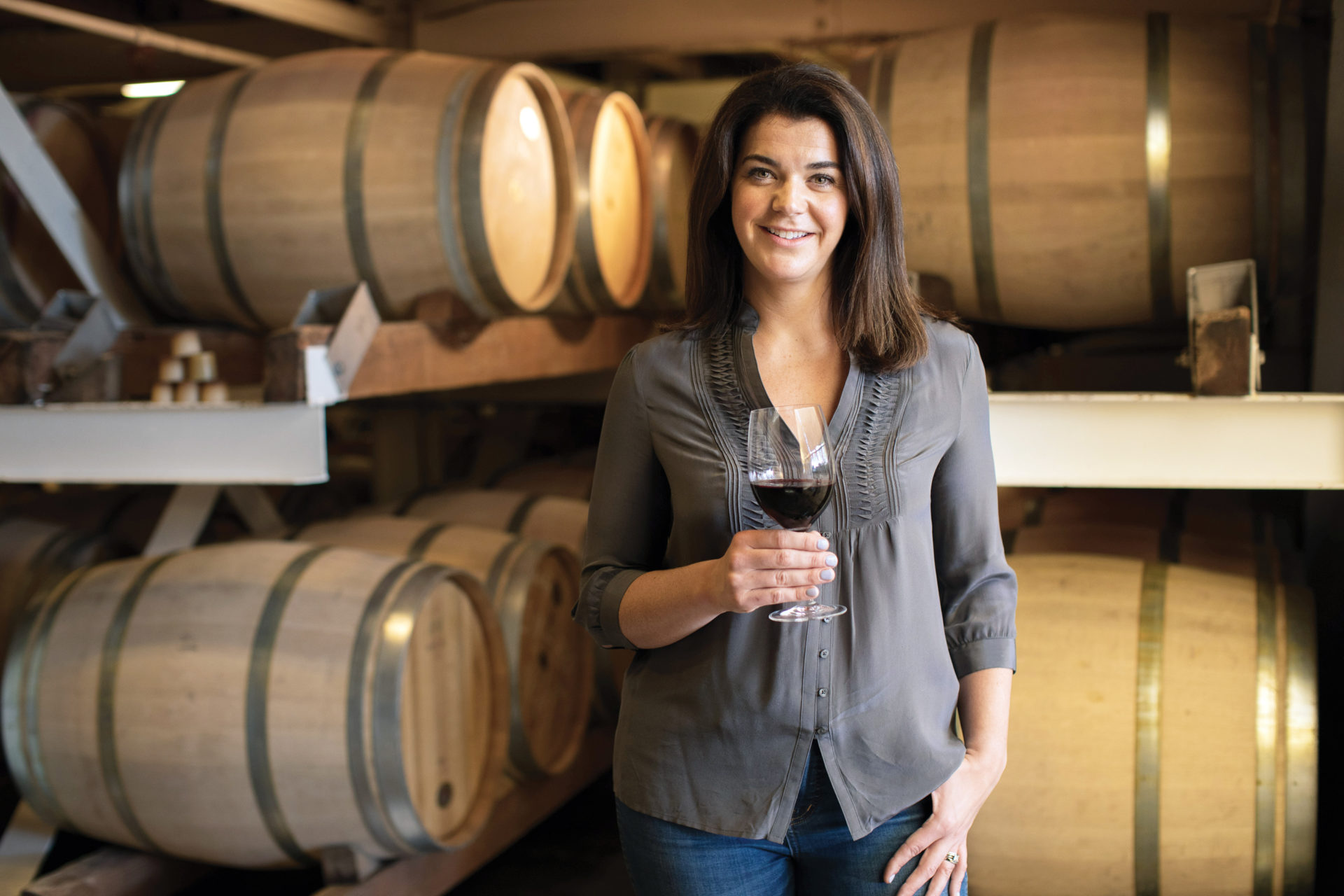 Winemaker Maggie Kruse holding a glass of red wine in front of brown wine barrels on white racks
