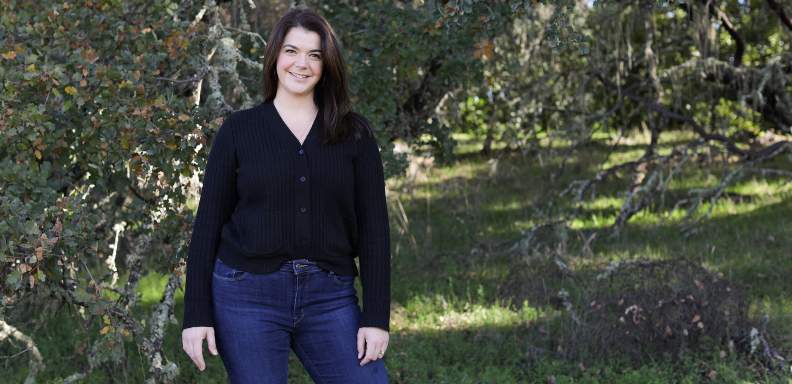 Woman in jeans and black shirt standing in a green meadow with trees behind her