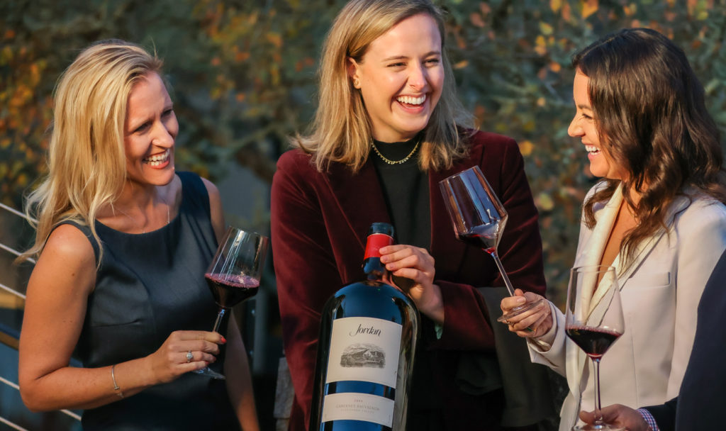 three women gathered around a large bottle of wine smiling