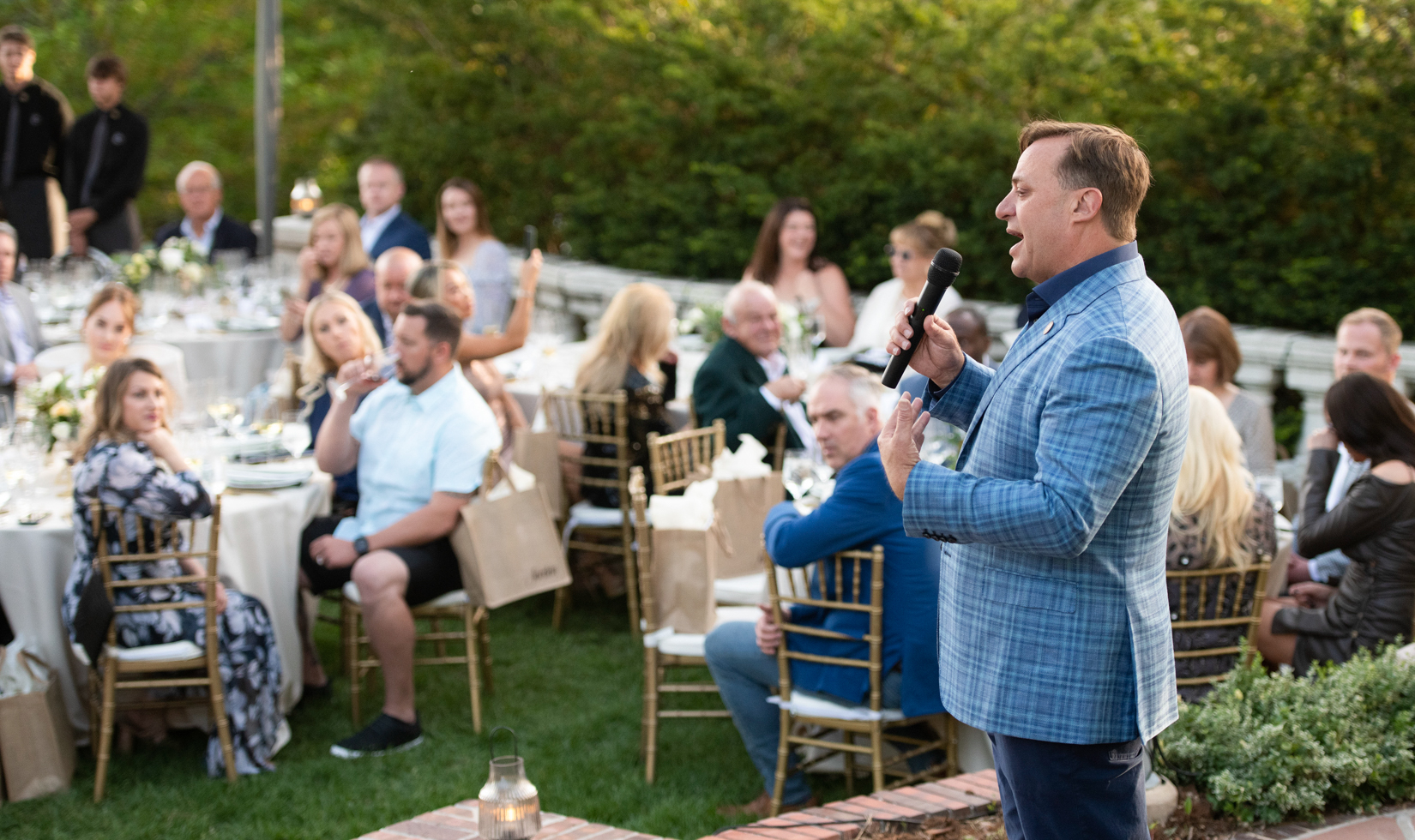 Man in blue blazer speaking to a group of seated guests at outdoor dinner 