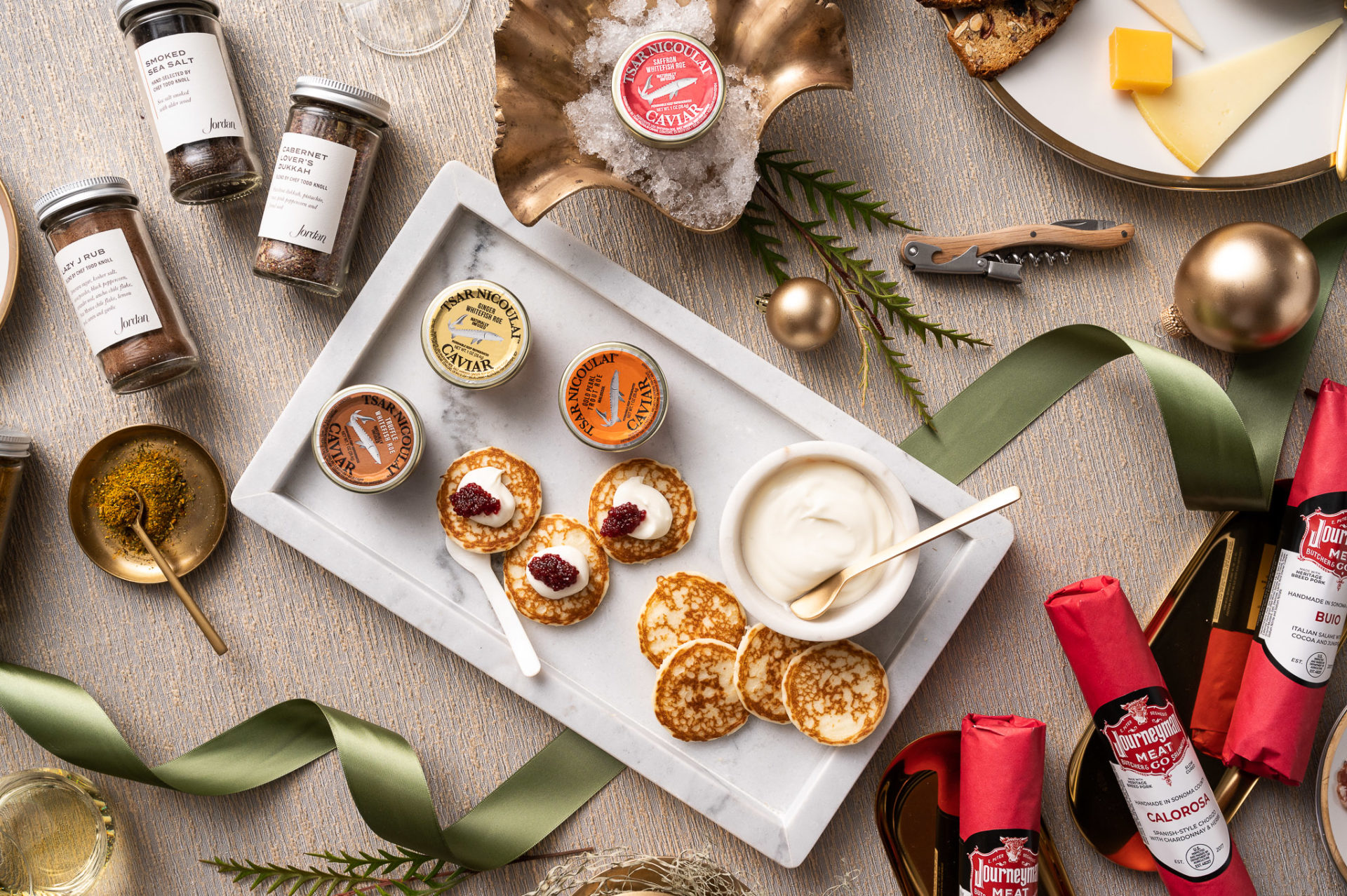 Marble tray with dishes of caviar and blinis with crème fraiche surrounded by jars of spices, salumi rolls, a corkscrew and cheese platter
