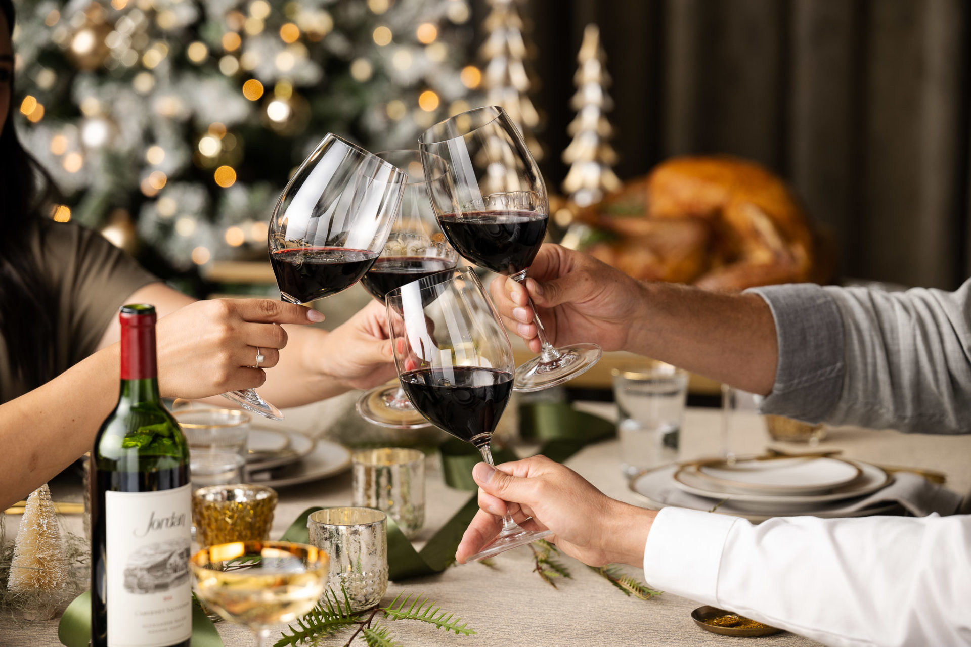 Three people toasting with glasses of cabernet seated around festive table decorated for the holidays with a lit Christmas tree and roast turkey in the background