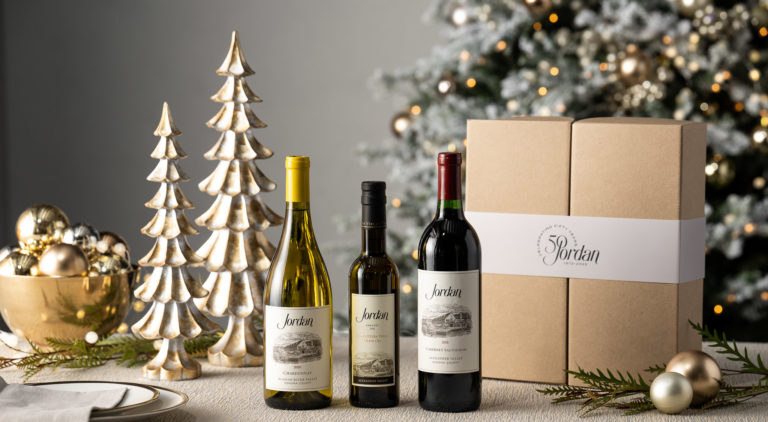 trio of chardonnay, olive oil and cabernet sauvignon bottles on table with gold ornaments and christmas tree with lights