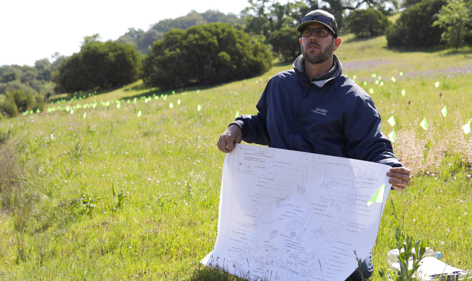 Farmer kneeling in a pollinator garden holding up renderings and plans