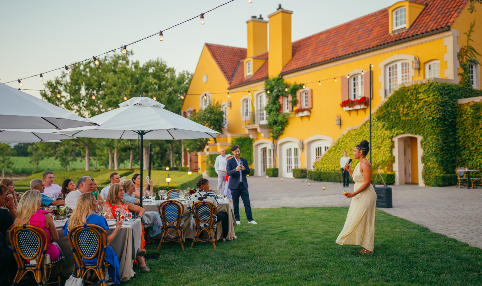 woman standing in front of seated guests with yellow chateau in the background.
