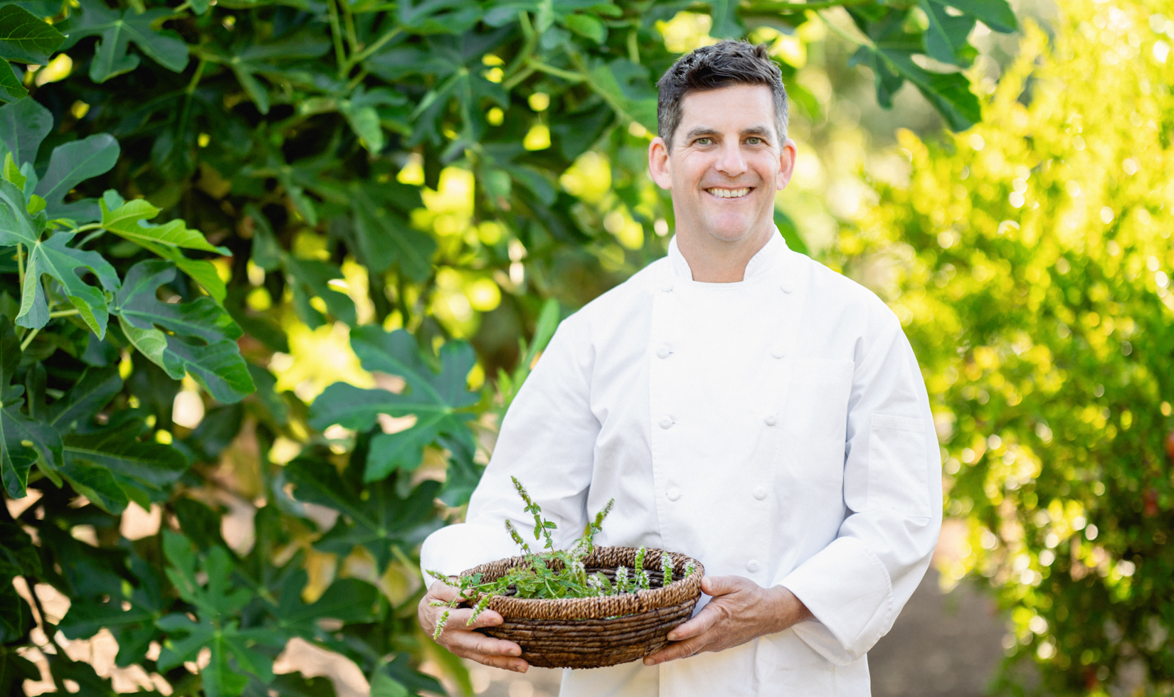 chef in white chef coat standing in green garden and holding basket of vegetables