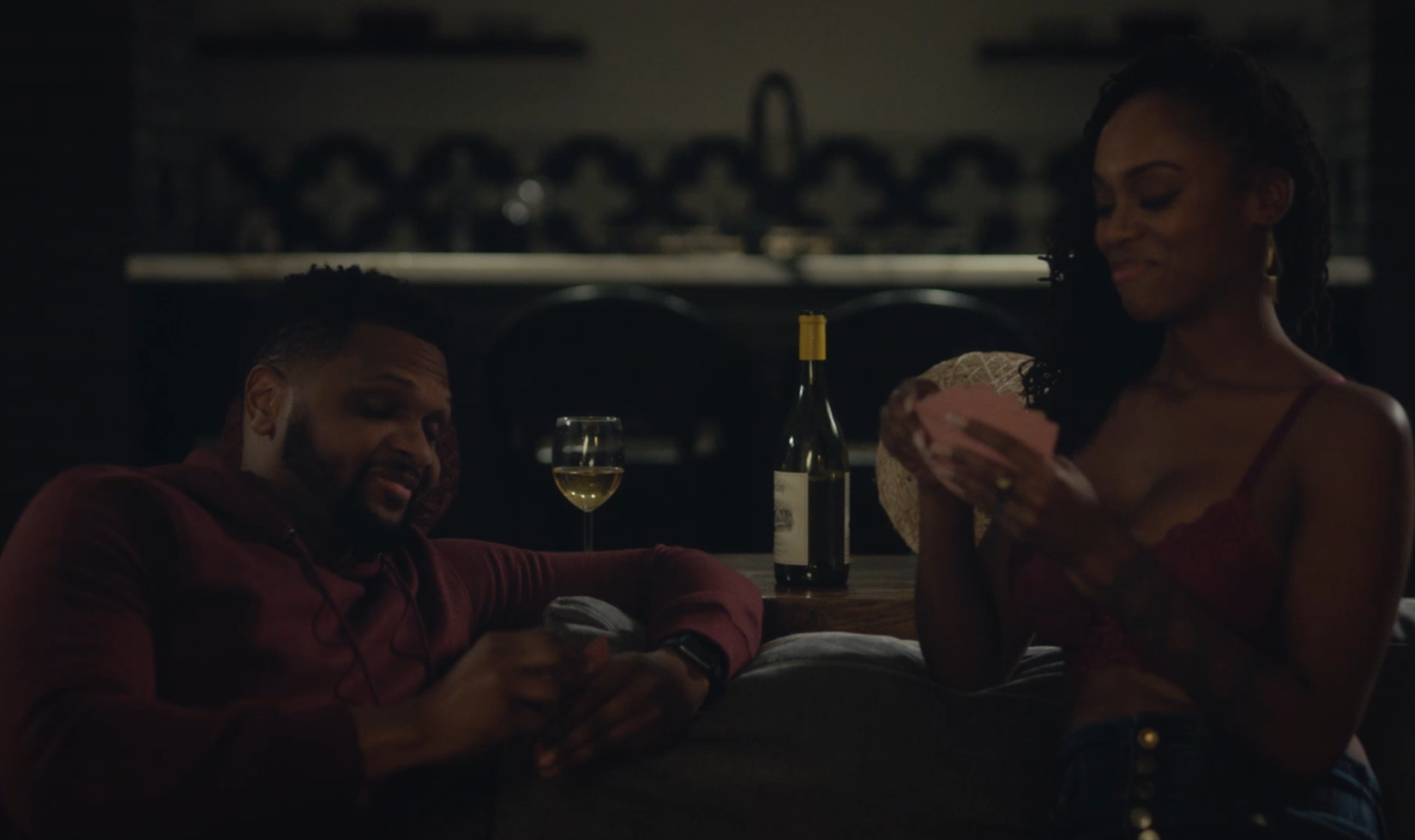 Screenshot from the TV series For the Love of Jason, showing a couple sitting a couch playing cards with a bottle of Jordan Chardonnay on the table behind them