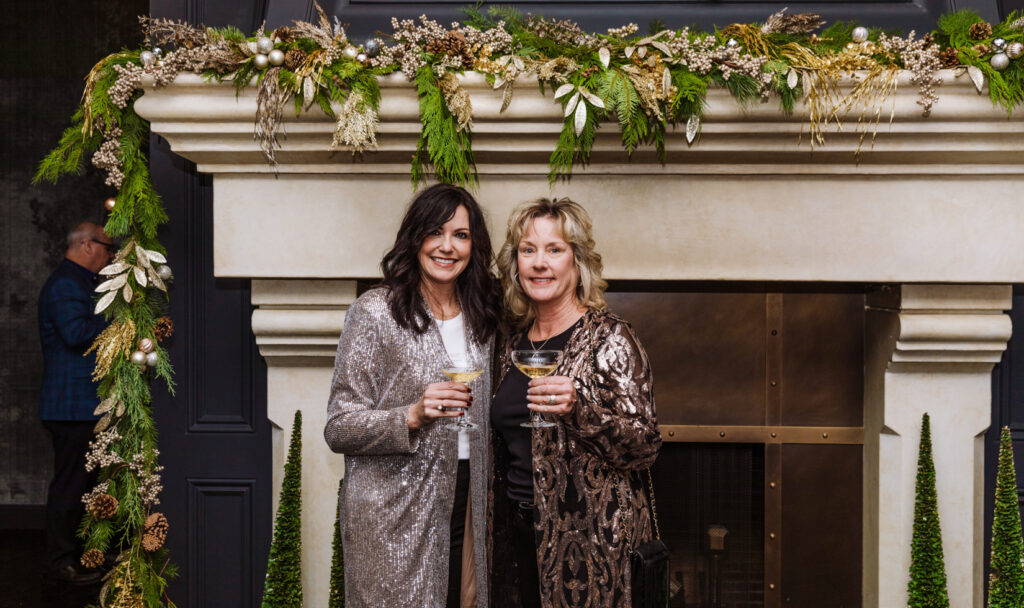 two woman with glasses of champagne in front of fireplace mantle decorated for christmas